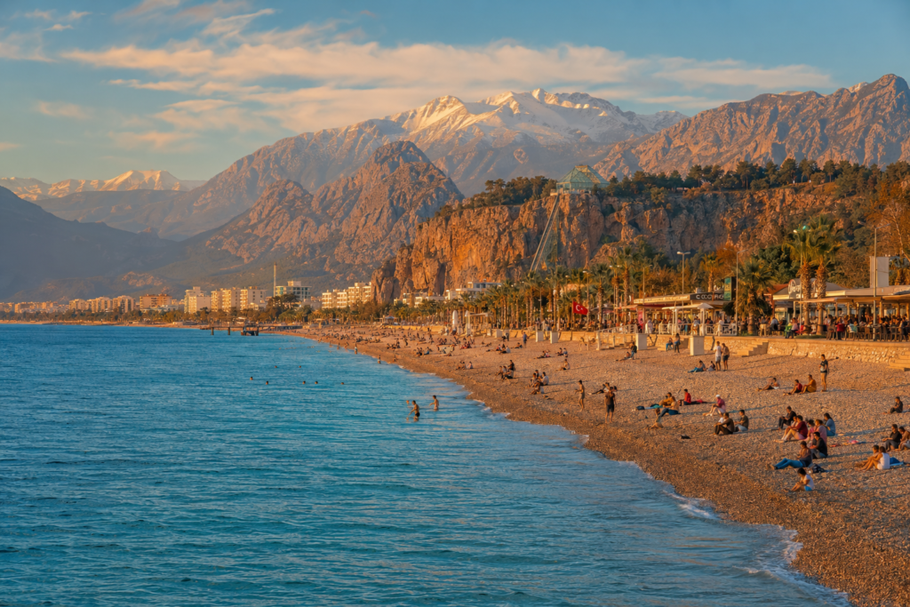 Konyaaltı Beach Antalya — crystal clear turquoise sea with dramatic Taurus Mountains backdrop and busy evening promenade atmosphere