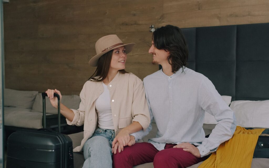 Couple sitting on a hotel bed with a suitcase before their trip